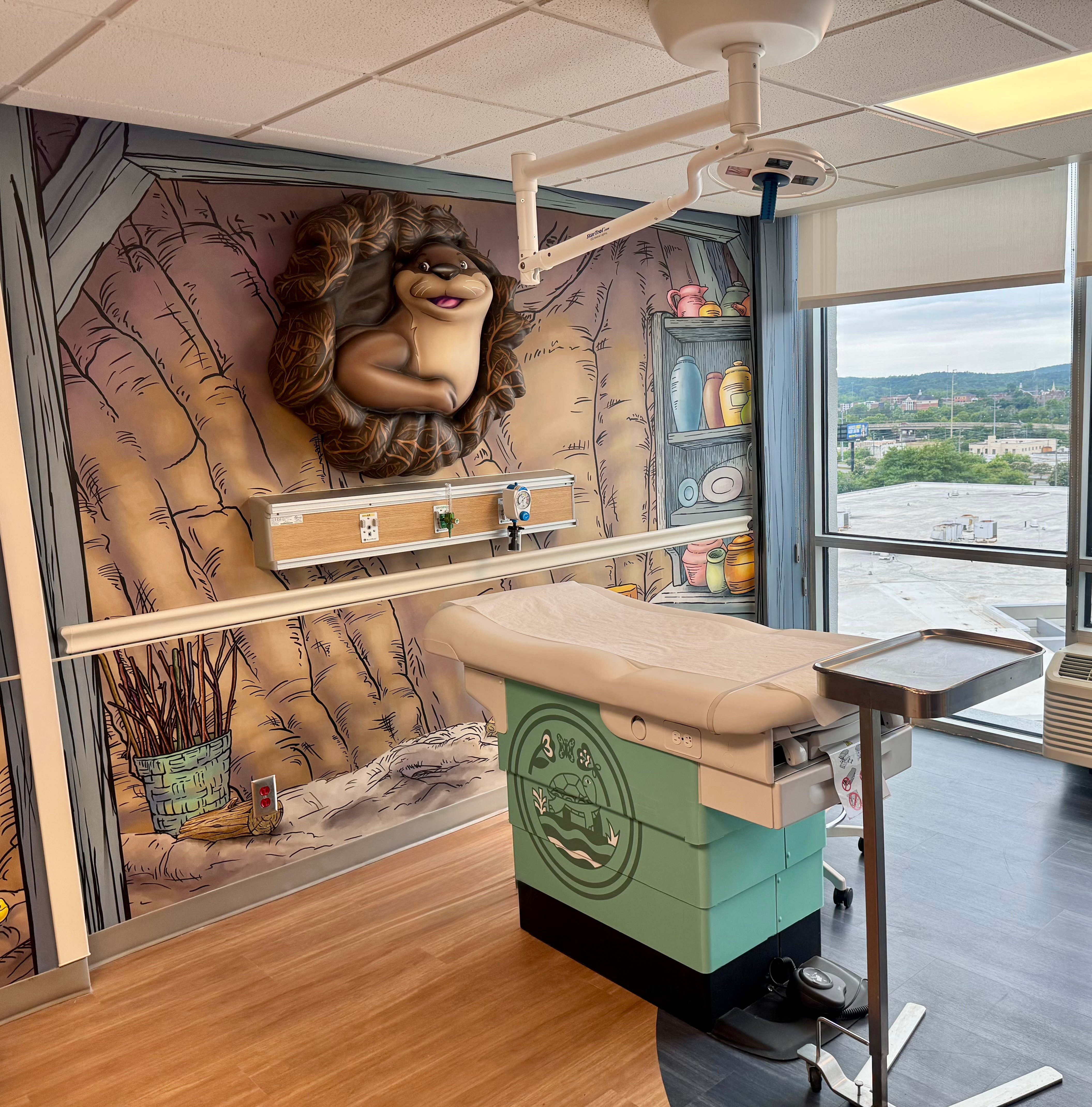 Pediatric treatment room at Bill & Olivia Amos Children's Hospital featuring Otto the otter, a custom dimensional wall sculpture, and immersive woodland-themed artwork designed to create a comforting environment for young patients