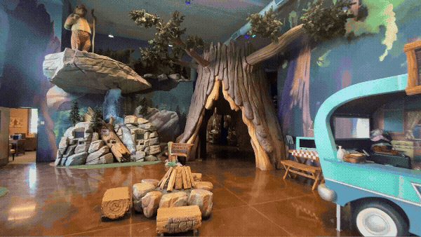 Camp-themed children’s play environment with a walk-through sculpted tree, rocky climbing features, campfire seating and interactive areas to explore.