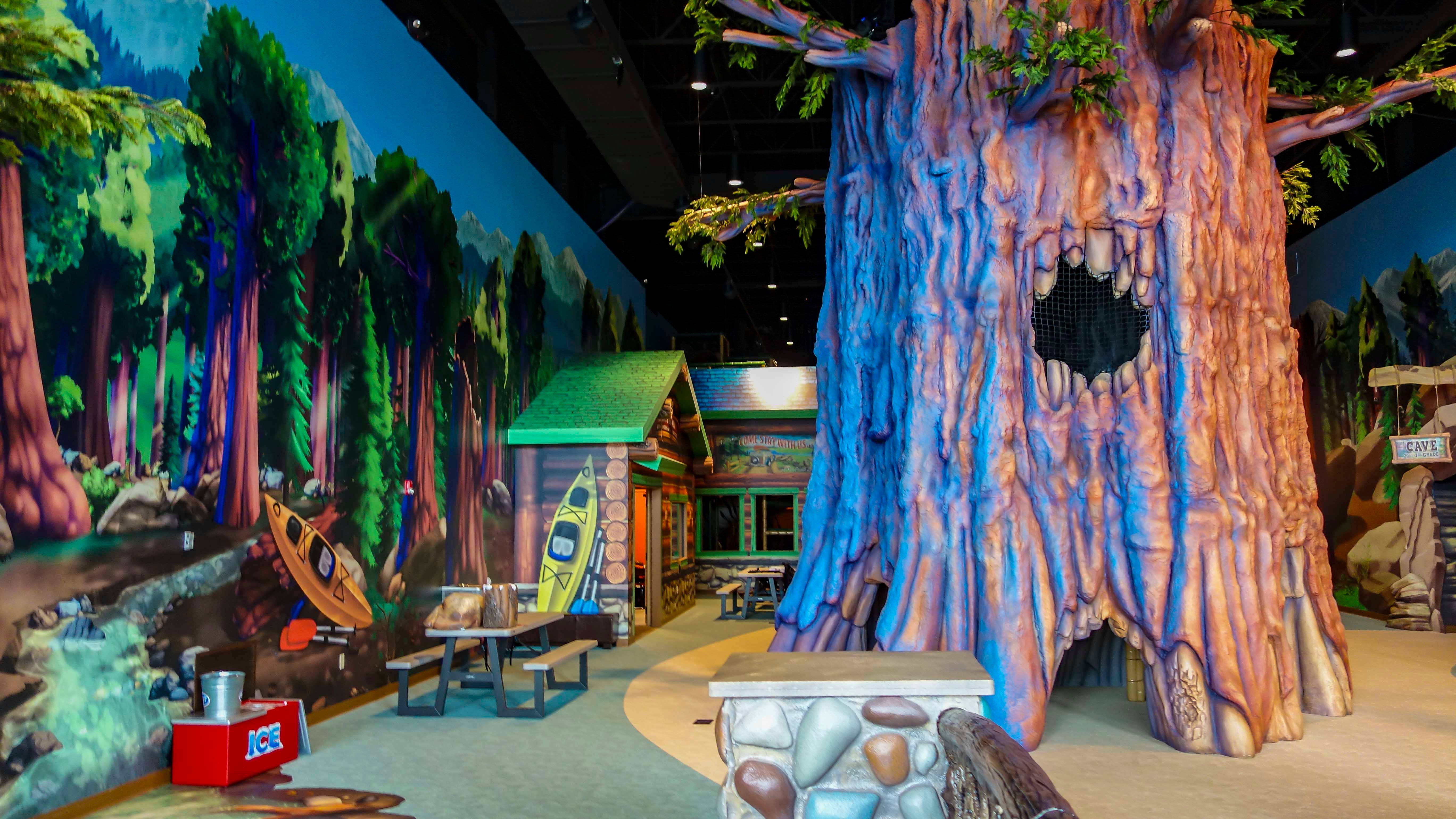 Immersive forest-themed children’s play space with a giant sculpted tree, cabin, stream mural and areas for kids to explore.