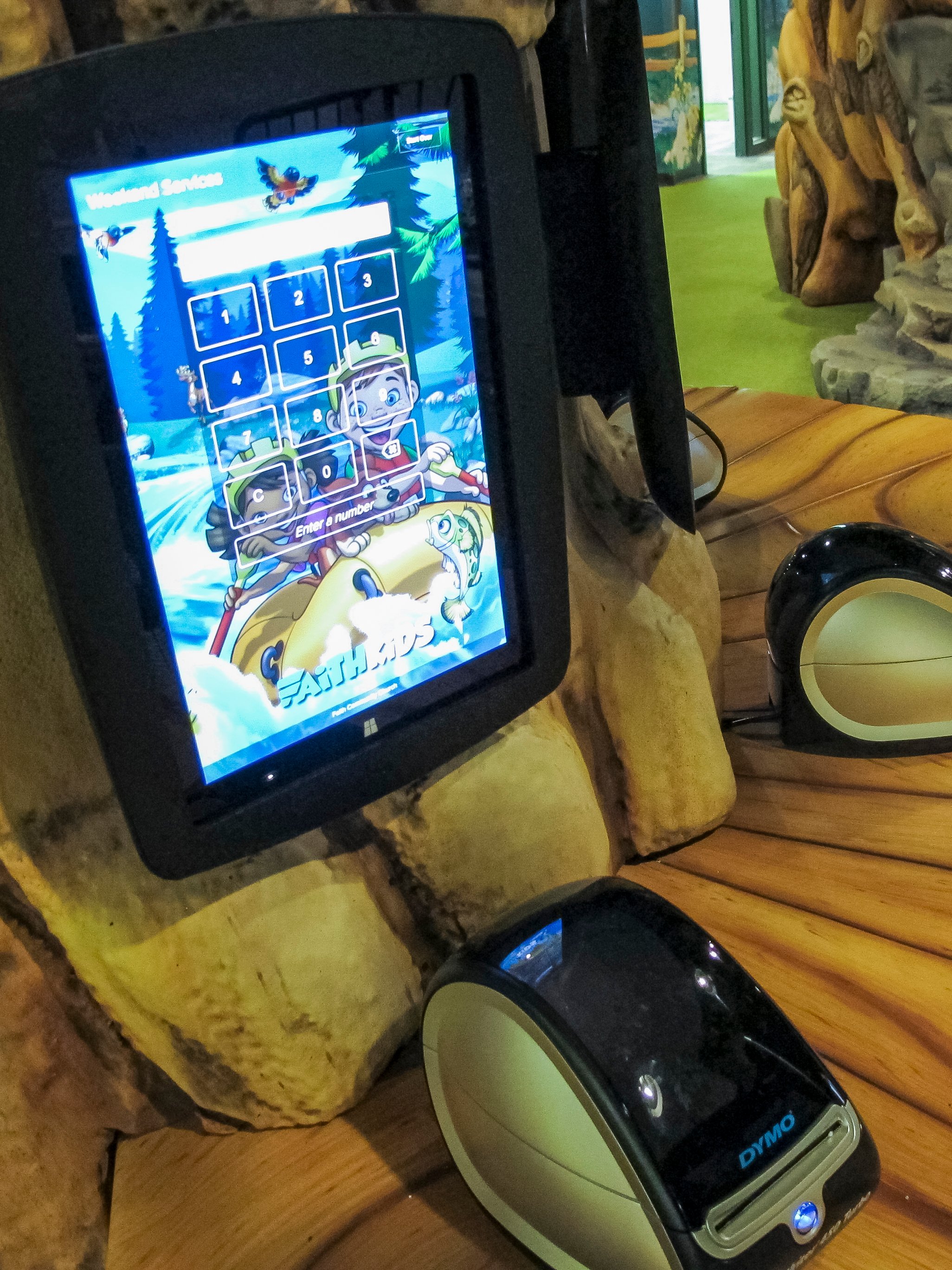 Integrated touchscreen check-in kiosk featuring custom camp-themed screen graphics and a label printer, extending the FaithKids theme into the digital check-in experience.