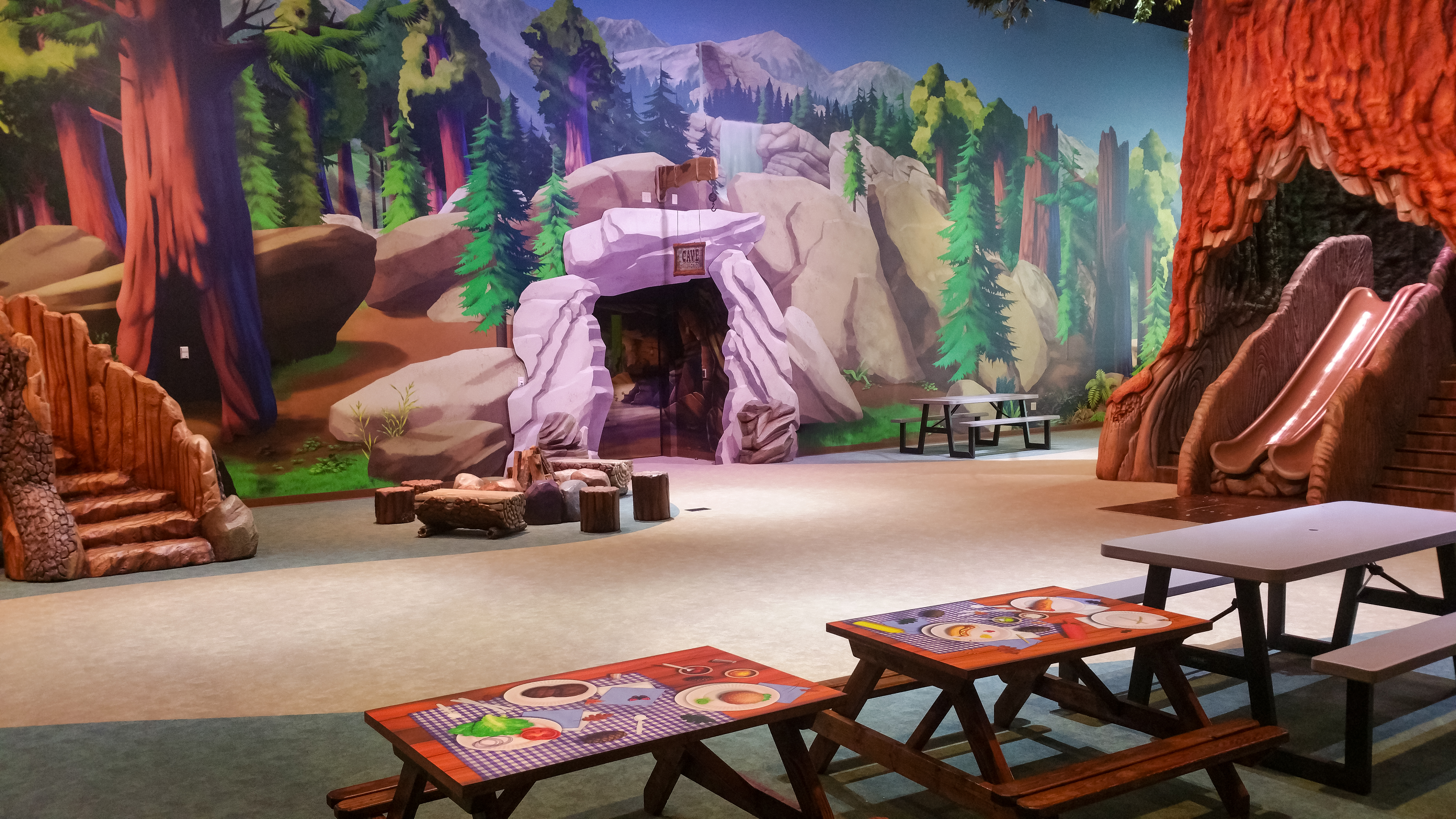 Forest-themed indoor play space featuring a giant tree slide, sculpted cave entrance, woodland murals, and picnic tables for families to gather and enjoy the space together.