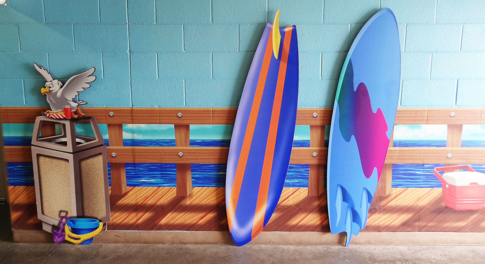 2D cutouts of surfboards nad waste can with bird and pail in front of fence wainscot in a Boardwalk Themed Space at Casas Church