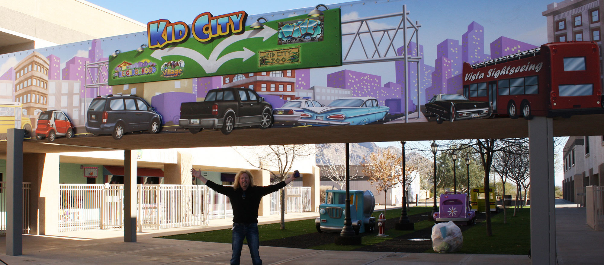 Bruce Barry underneath a large outdoor mural of a Toon Town Big City skyline with sign reading "Kid City" at Casas Church
