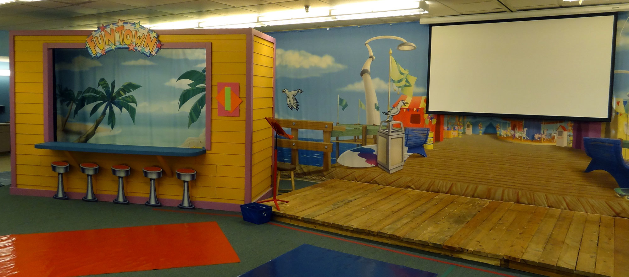 Wooden stage and Boardwalk scene mural plus a 3D puppet stage shack with a beach scene background at a Beach and Boardwalk Stage Themed Environment at Casas Church
