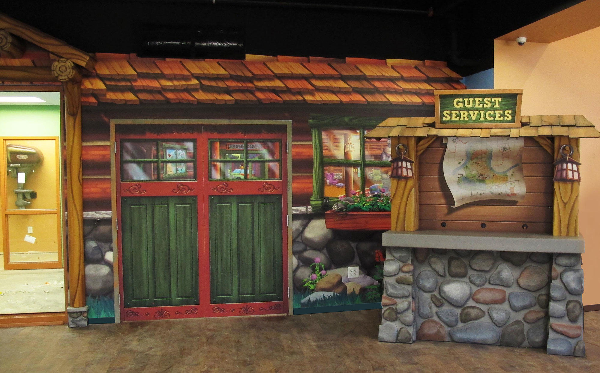 Camping Themed Facade of a lodge with double doors and a window plus a 3D sculpted rock and wood check in kiosk with sign reading "Guest Services" at a Church