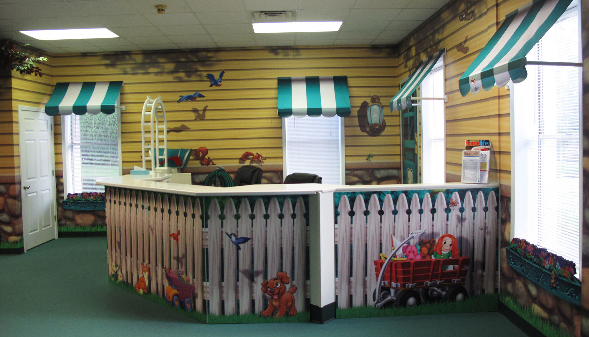 Neighborhood Themed Check In Area with white picket fence desk wrap and yellow house murals with green and white awnings over windows at a Church