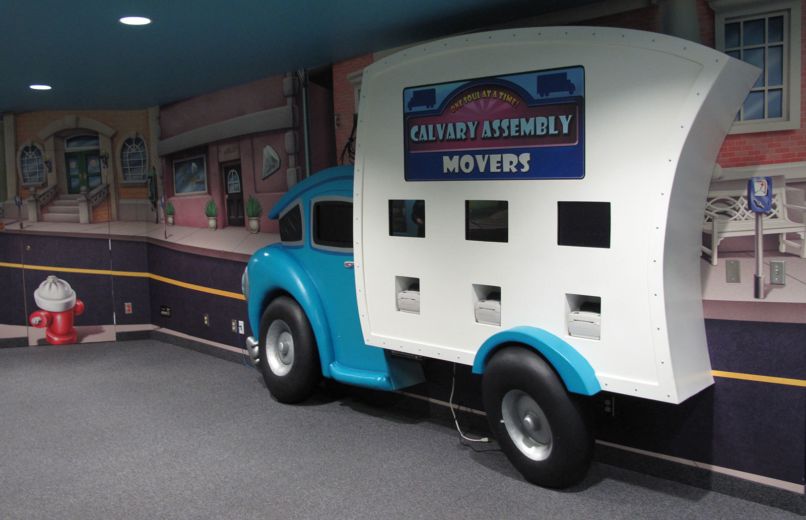 3D relief sculpted blue and white delivery truck with 3 Check-In Kiosks in a Toon Town Themed Space with full wall mural of a downtown scene.
