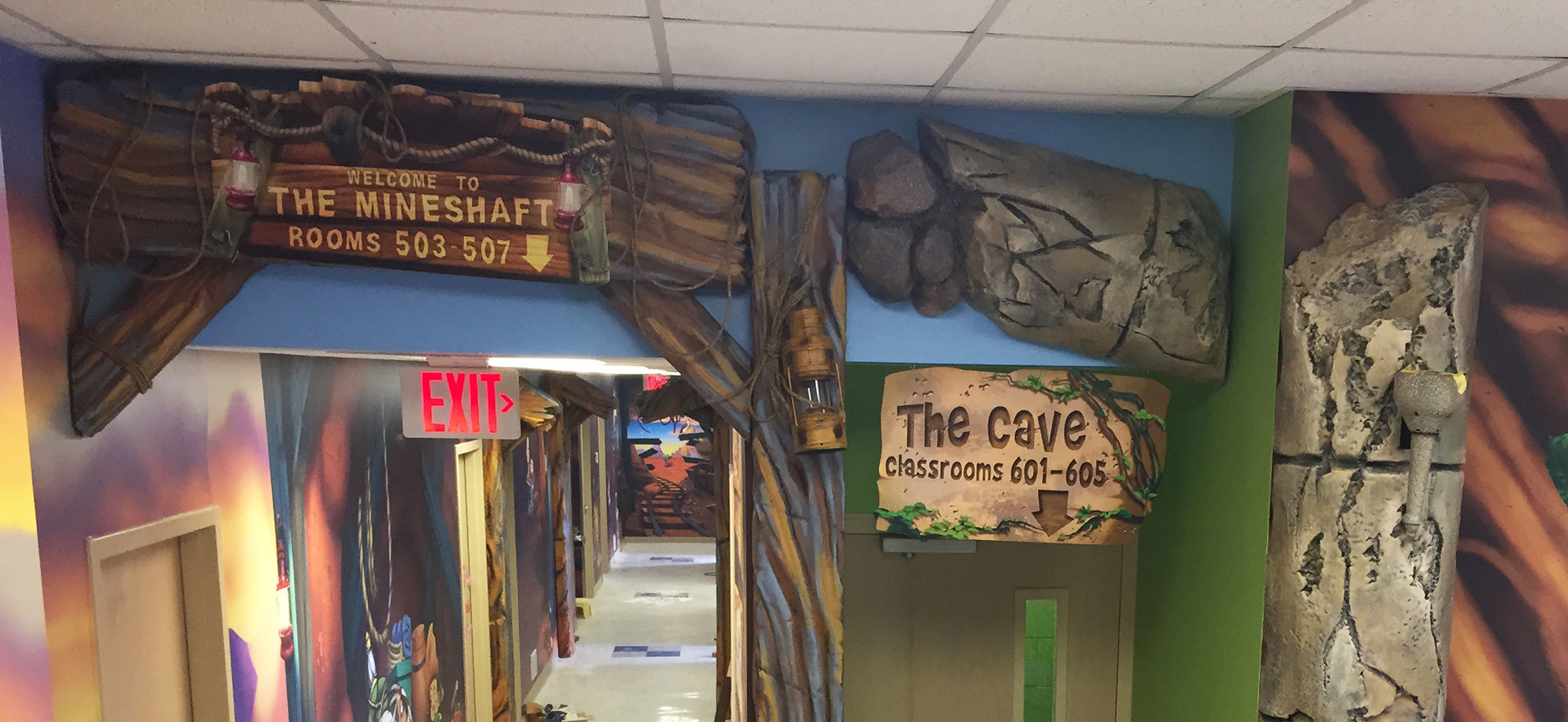 2D cutout sign reading "The Mineshaft Rooms 503-507" plus sculpted rocks and a asign reading "The Cave Classrooms 601-605" and murals depicting a mine scene.