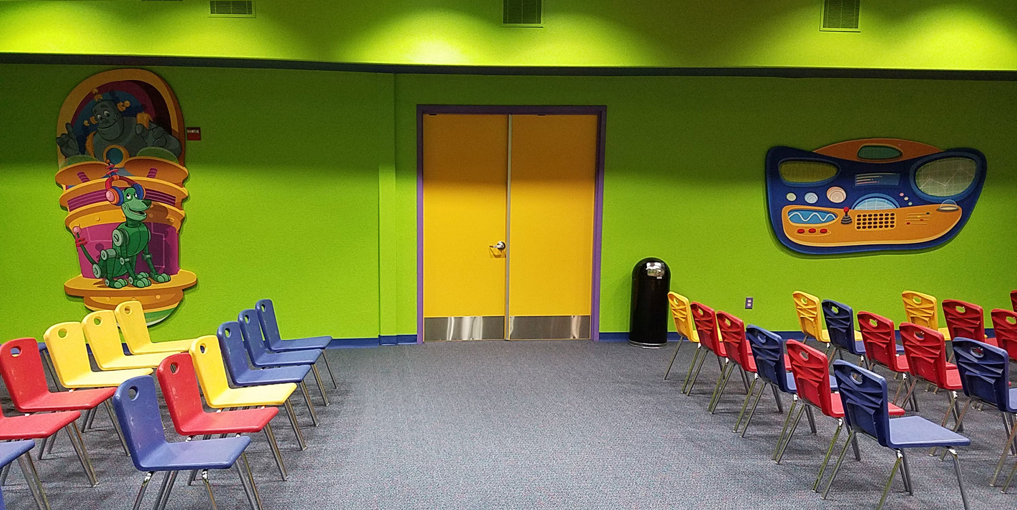 Multi colored chairs in a green room with yellow double doors and 2 cut out sound panels; one is a robot DJ with robot dog the other is a wacky control panel in an Outer Space Future World Themed room