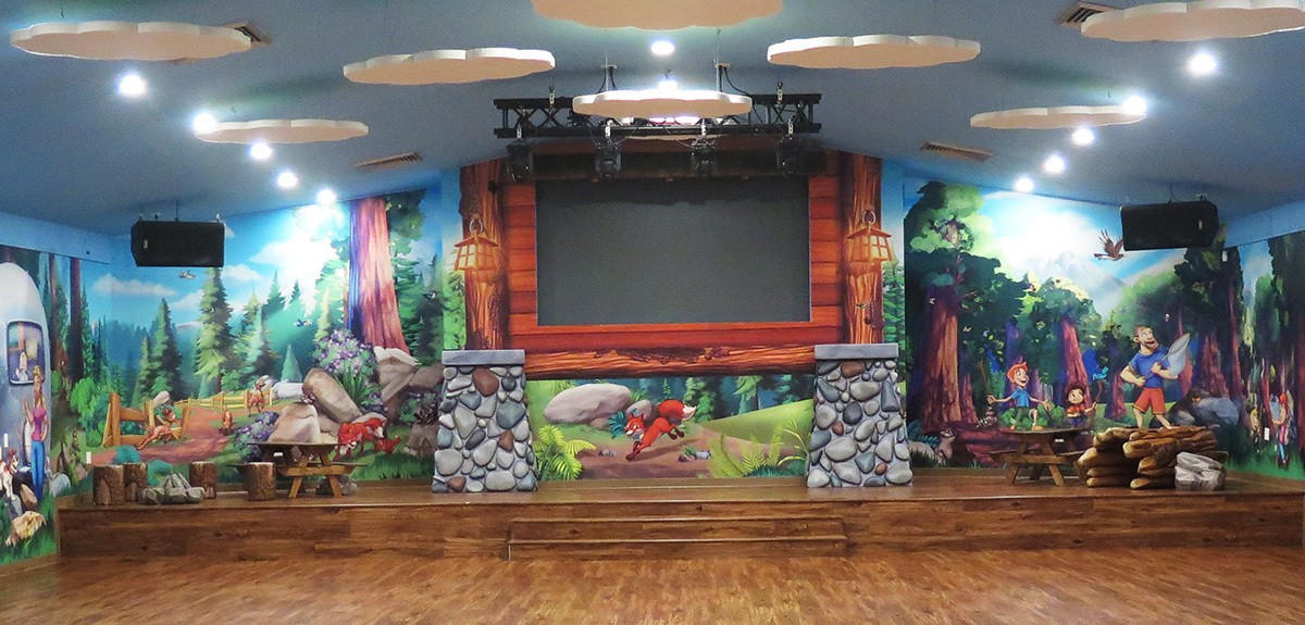 Camping/Lodge themed stage with sculpted rocks, log, picnic tables and forest mural background.
