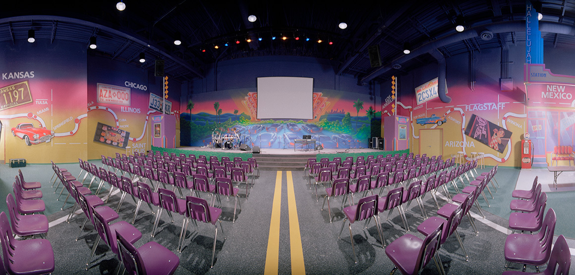 Large theater with a Route 66 themed wall covering including a stage backdrop plus rows of purple chairs.