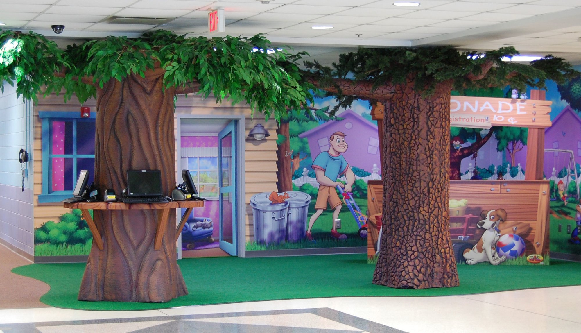 2 3D sculpted trees as check in desks, a faux grass floor plus a wall mural neighborhood scene with a yellow house, a man mowing grass and a lemonade stand with a dog in front.