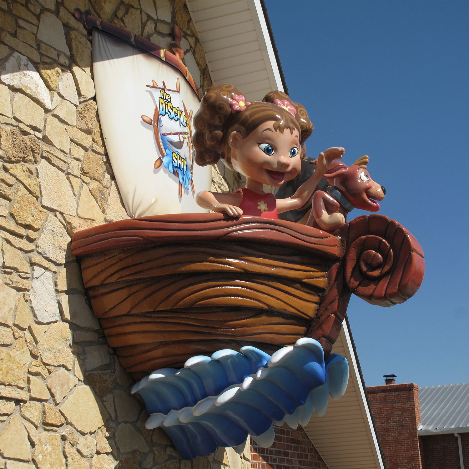 Outdoor 3D Sculpted Sign of Children in a Sailboat atop a wave jutting out on a high outdoor stone wall of a church