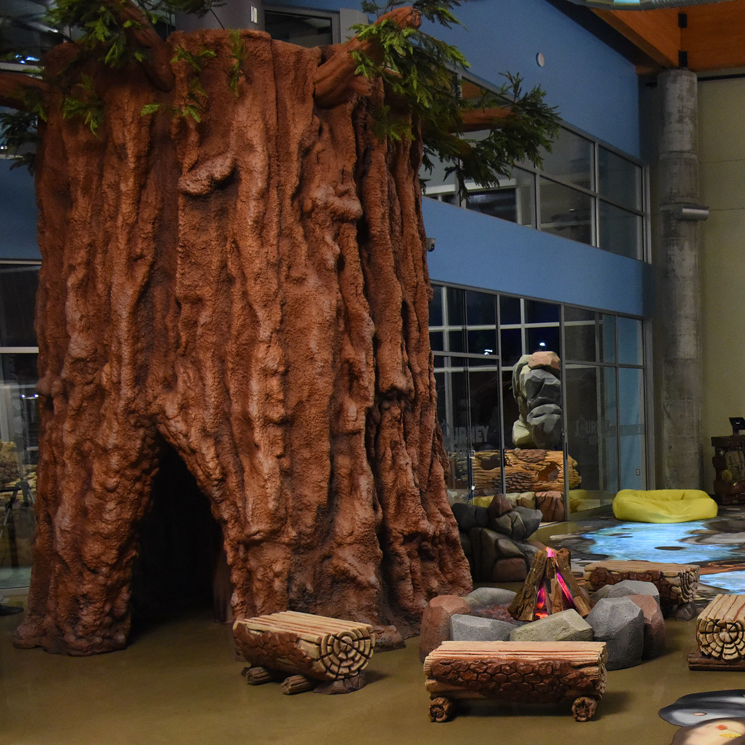 3D sculpted tree with crawl through, 3D fallen logs as benches, a fire pit, an animated stream and a yellow life raft in a Camping Themed Space at Abundant Life Church