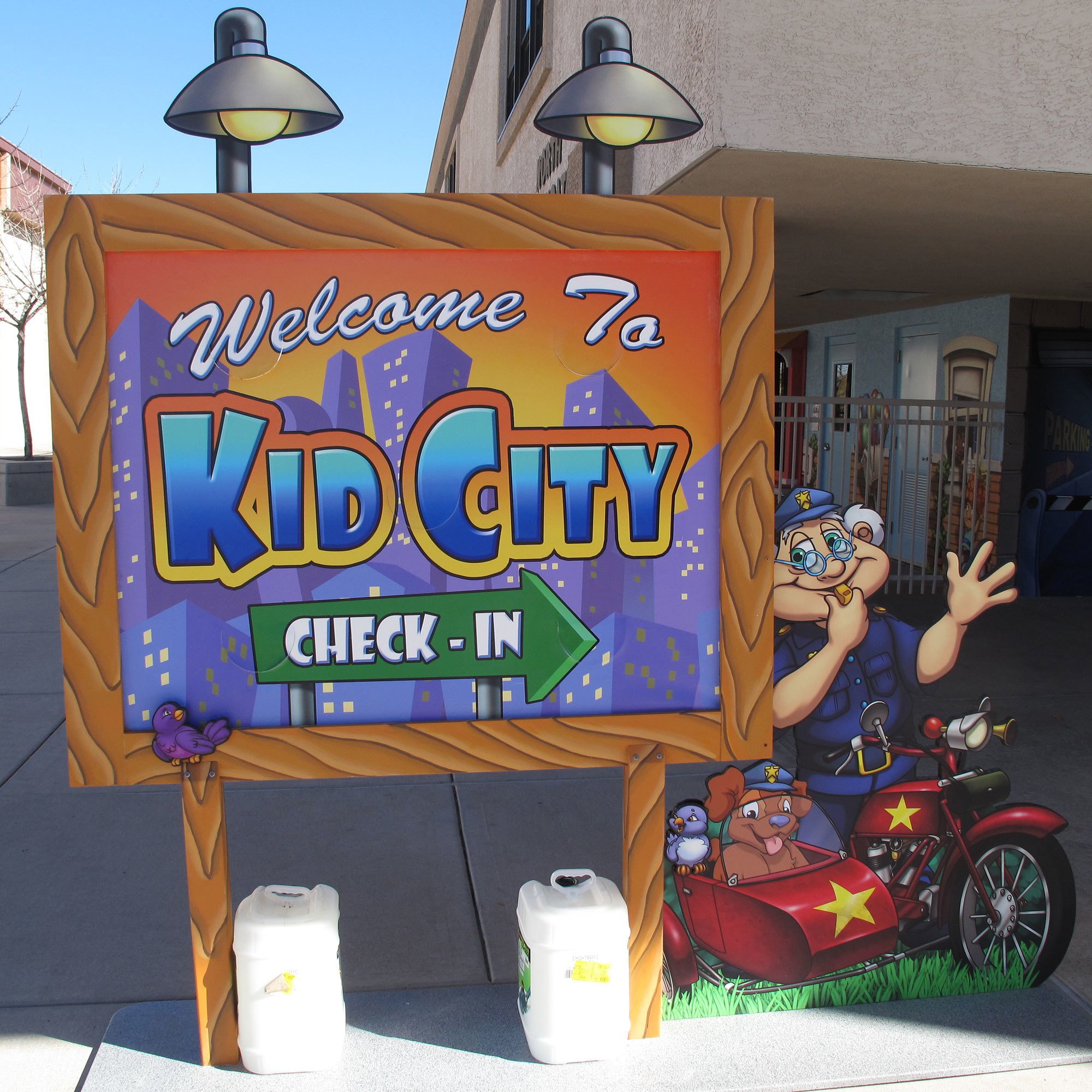 2D cutout direction sign reading "Welcome To Kid City Check In" with a green arrow plus a 2D cutout of a cartoon cop on a motorbike outside Casas Church
