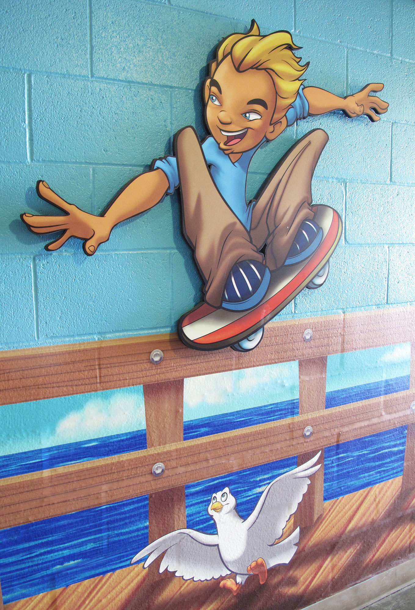 2D cutout of blonde boy on skateboard atop wooden fence with white seagull below in a Boardwalk Themed Space at Casas Church