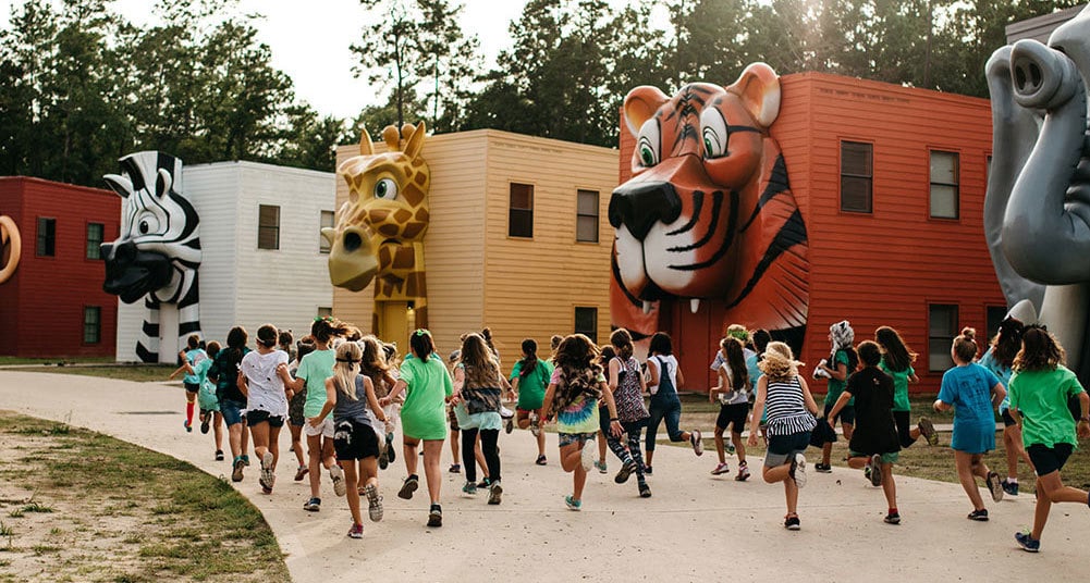 Group of Teen Girls Running in Front of Giant Animal Building Facades on Camp Cabins