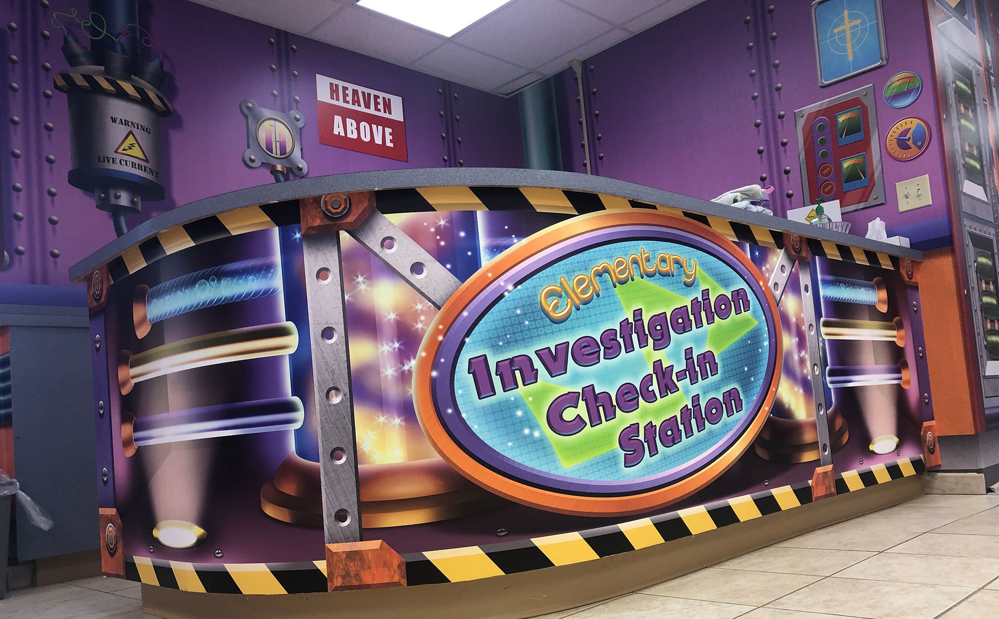 Industrial Science Lab Factory Themed Wall Covering and wrapped desk with sign reading "Elementary Investigation Check In Station" at London Bridge Baptist Church