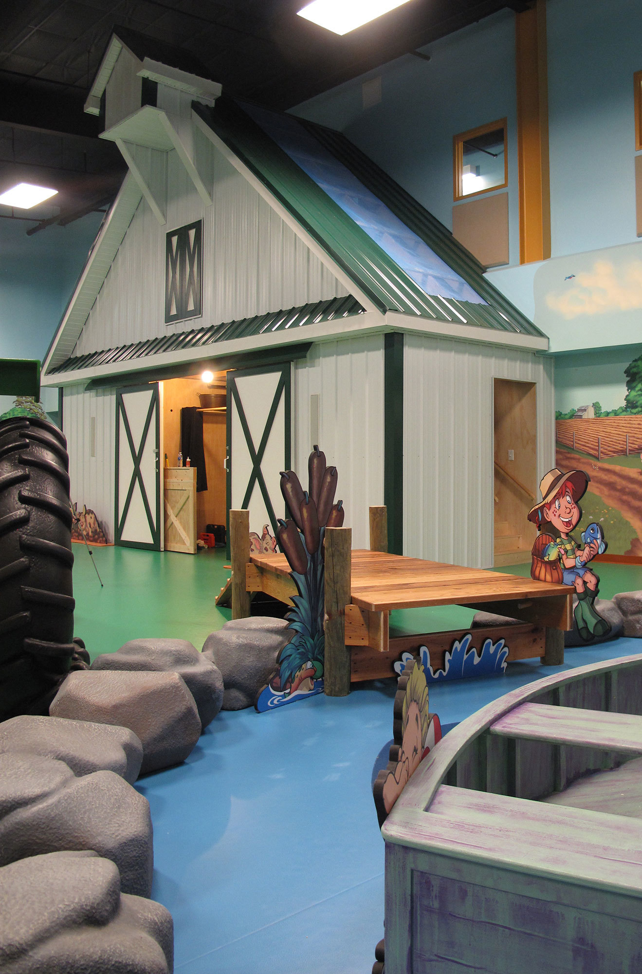 White Barn with Green roof, 3D sculpted rocks surround painted lake with 2D cutouts of kids, a dock and boat in a Farm Themed Space at Hardin Baptist Church