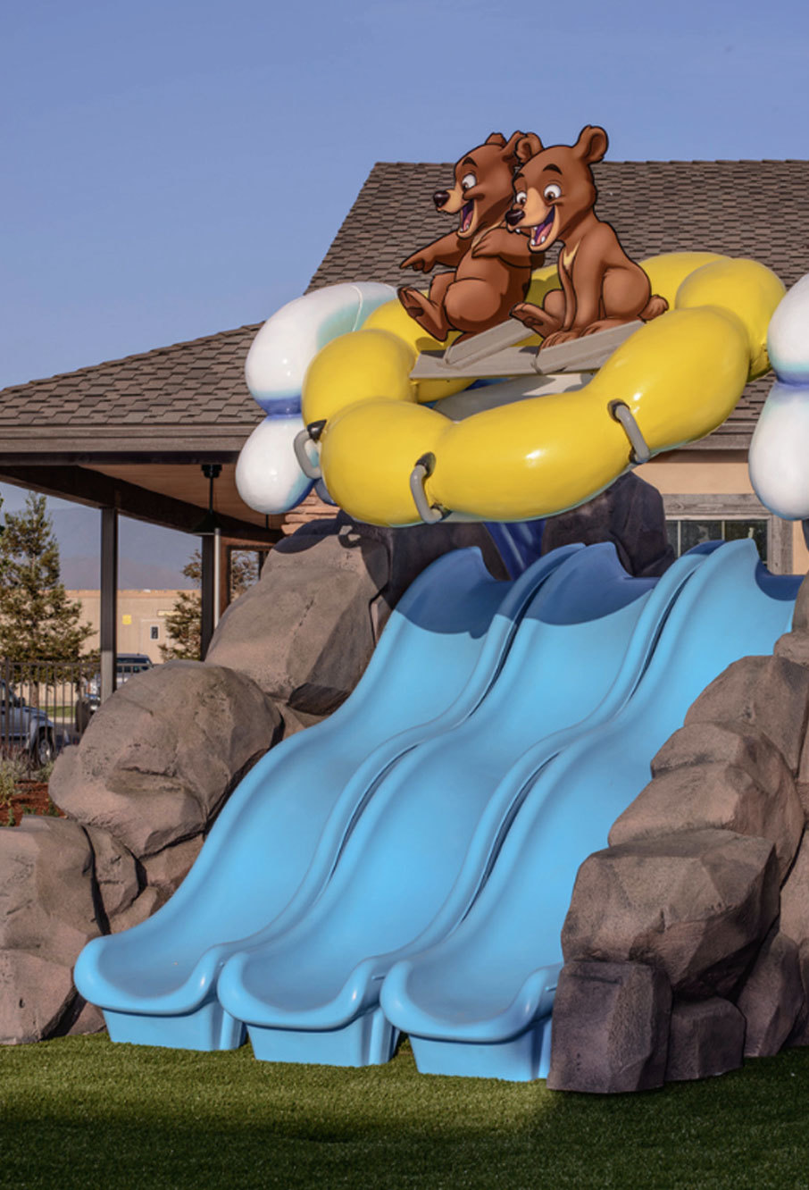 Triple blue slides on a 3D sculpted rocks base with bears in a yellow raft riding a wave as an outdoor play feature in a Camping Themed Environment at Canyon Hills Church