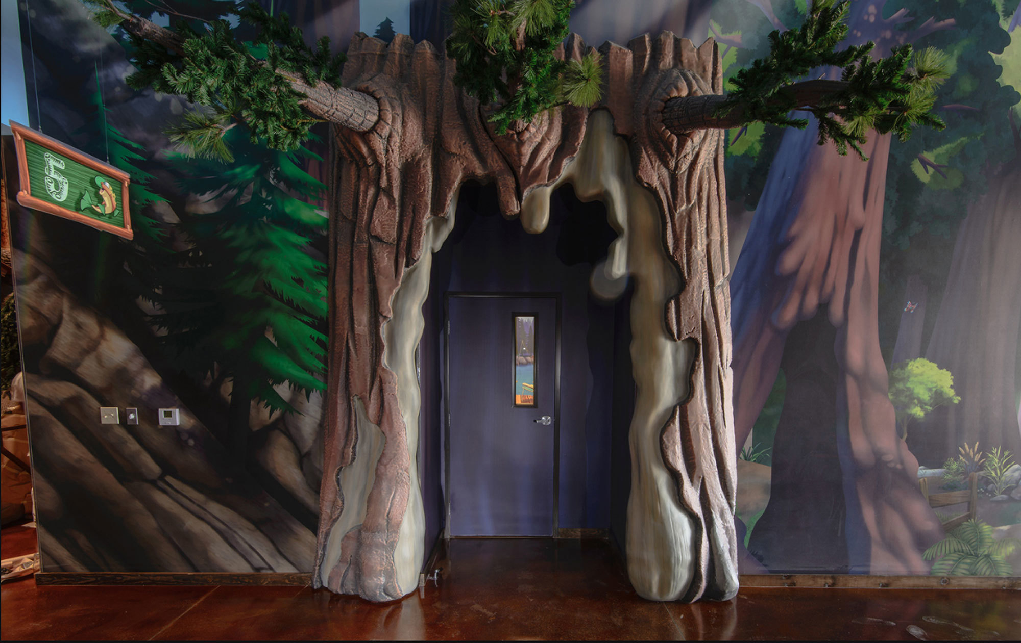 Sculpted 3D relief tree surrounding a doorway with Forest scene wall murals and 2D classroom signs in a Camping Themed Environment at Canyon Hills Church