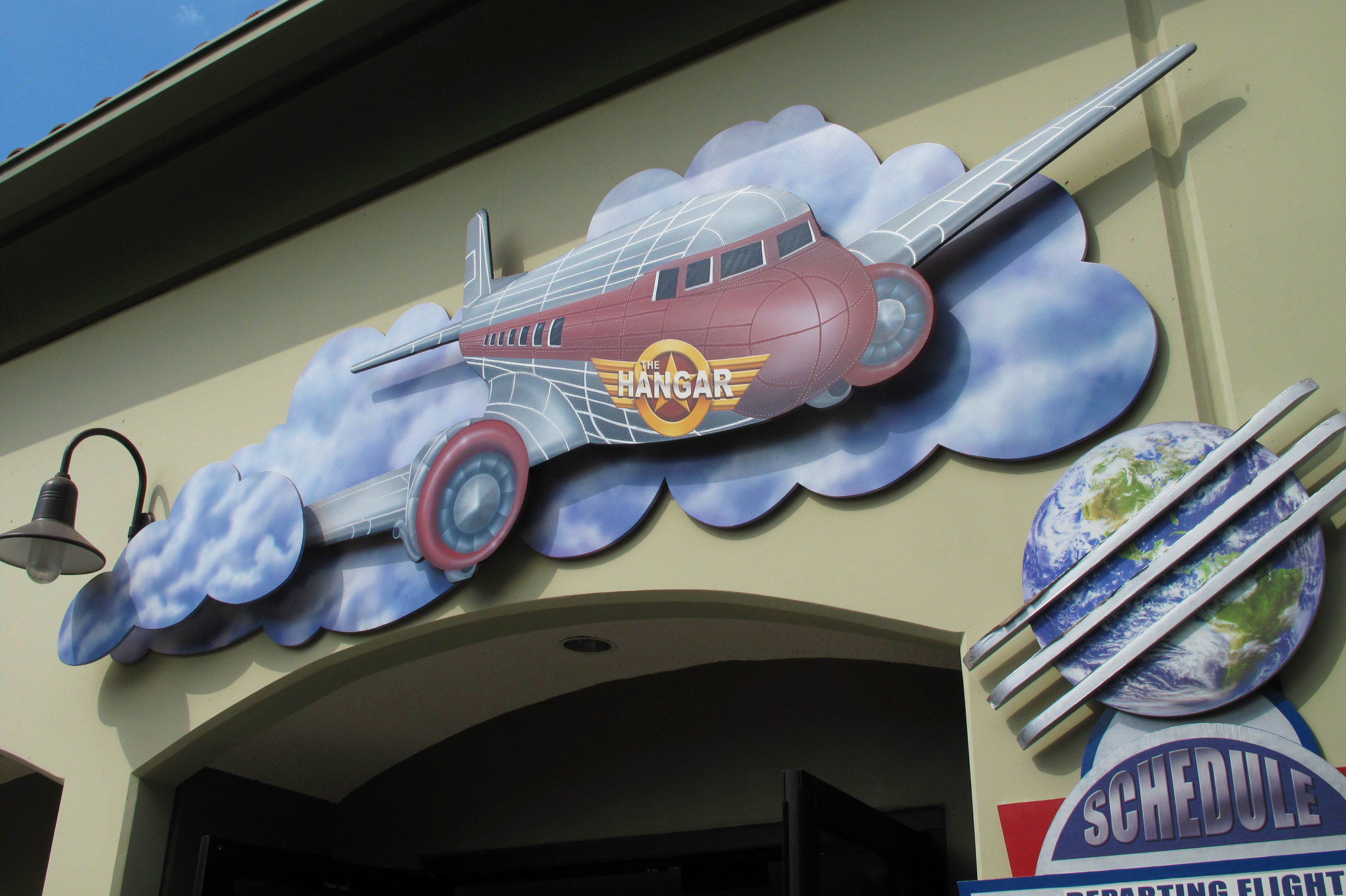 Outdoor 2D Sign with clouds and an airplane with emblem reading "The Hangar" above an entryway