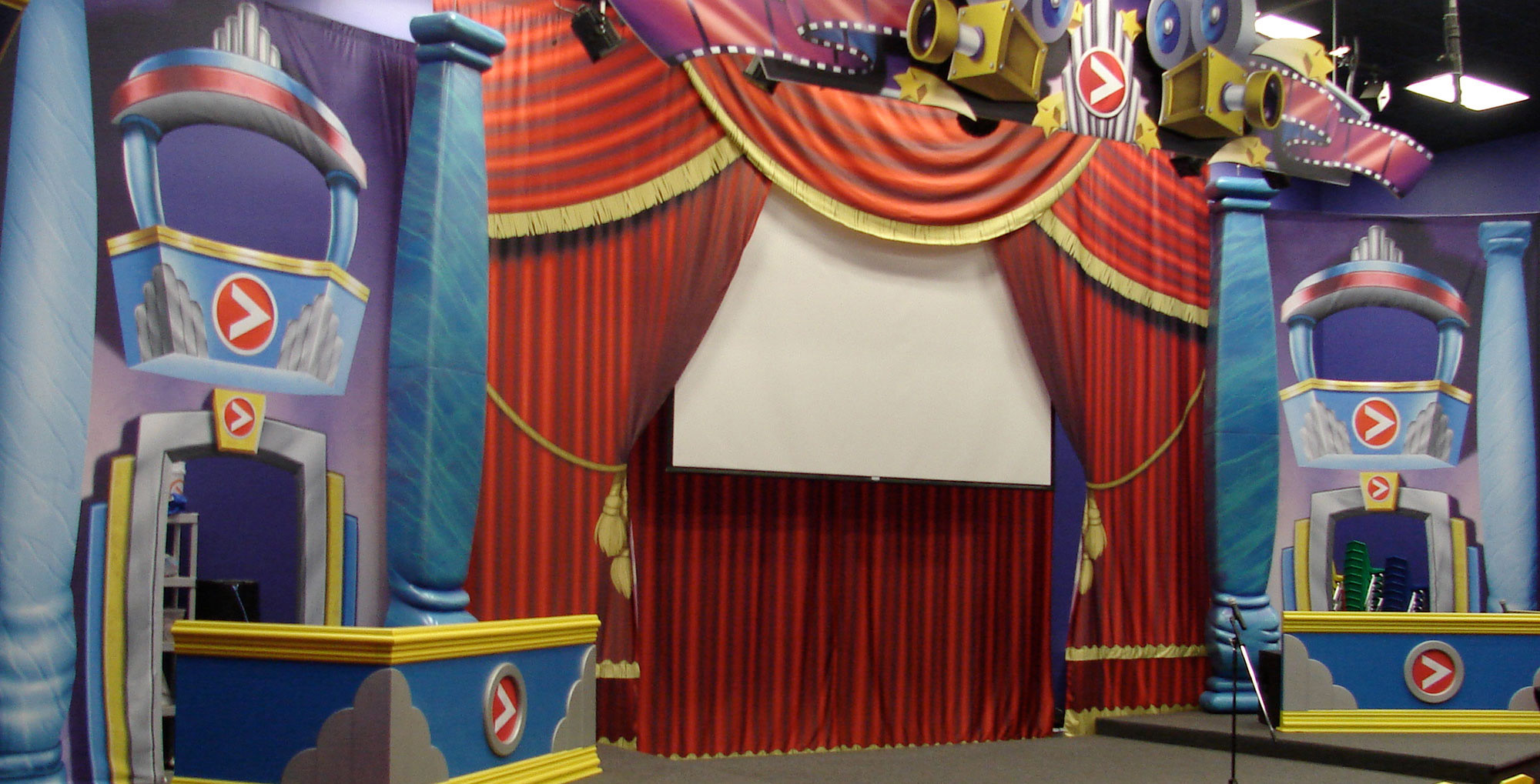 A large themed stage with Red curtains with gold fringe, blue sculpted pillars, side entries and a large 2D Back Lot Studio themed Sign.