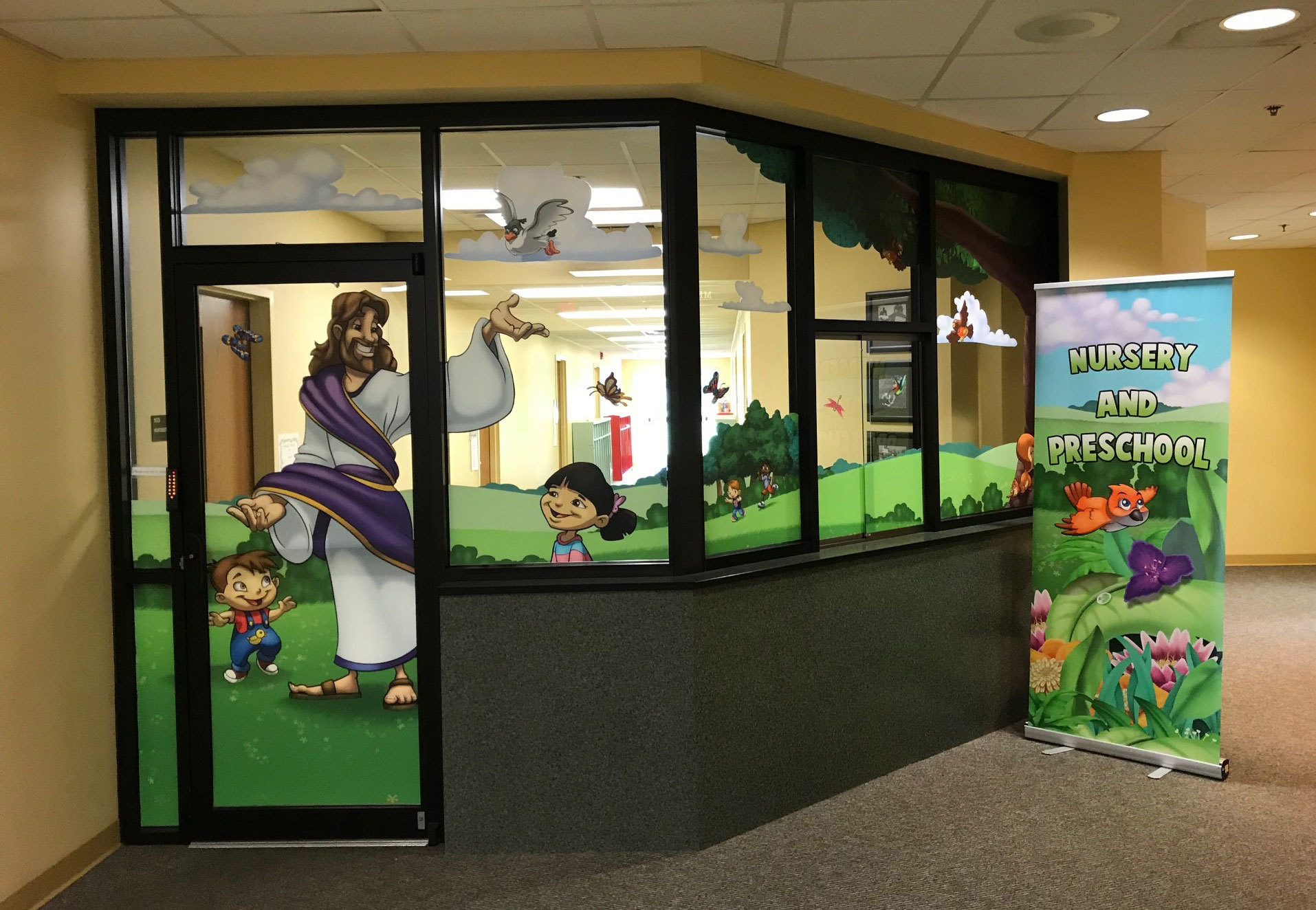 Biblical Window Vinyl depicting Jesus and children in a meadow and Pop Up Banner sign with flowers and bird reading "Nursery and Preschool" at a Church Office